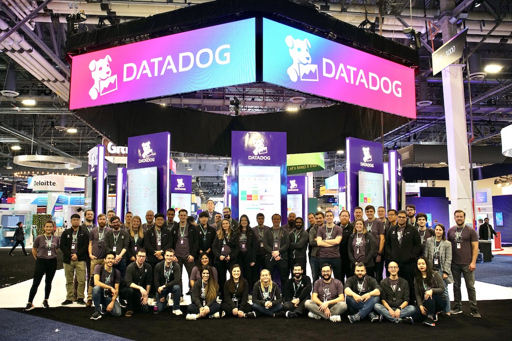 A large group of Datadog team members gathers underneath a large neon sign with the company logo.