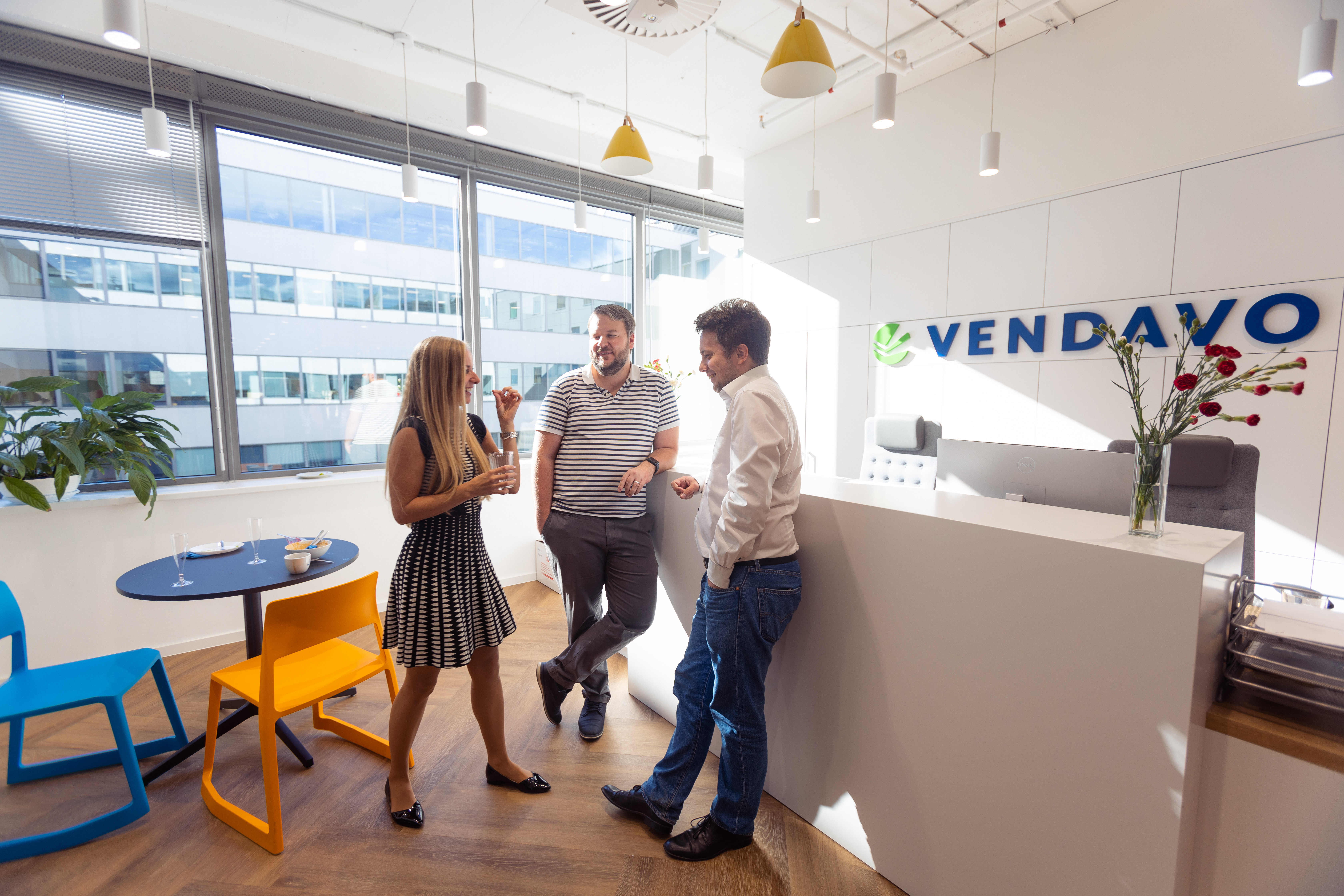 Vendavo's office. Two men and a woman are speaking while the men are leaning against a white counter. On the counter is a vase of roses. Next to the woman is a table and two brightly-colored chairs.