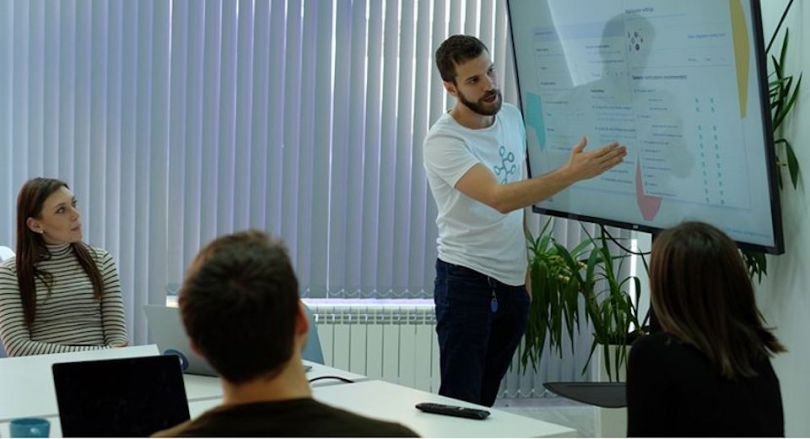 A man at a whiteboard, with several people at a table watching him.