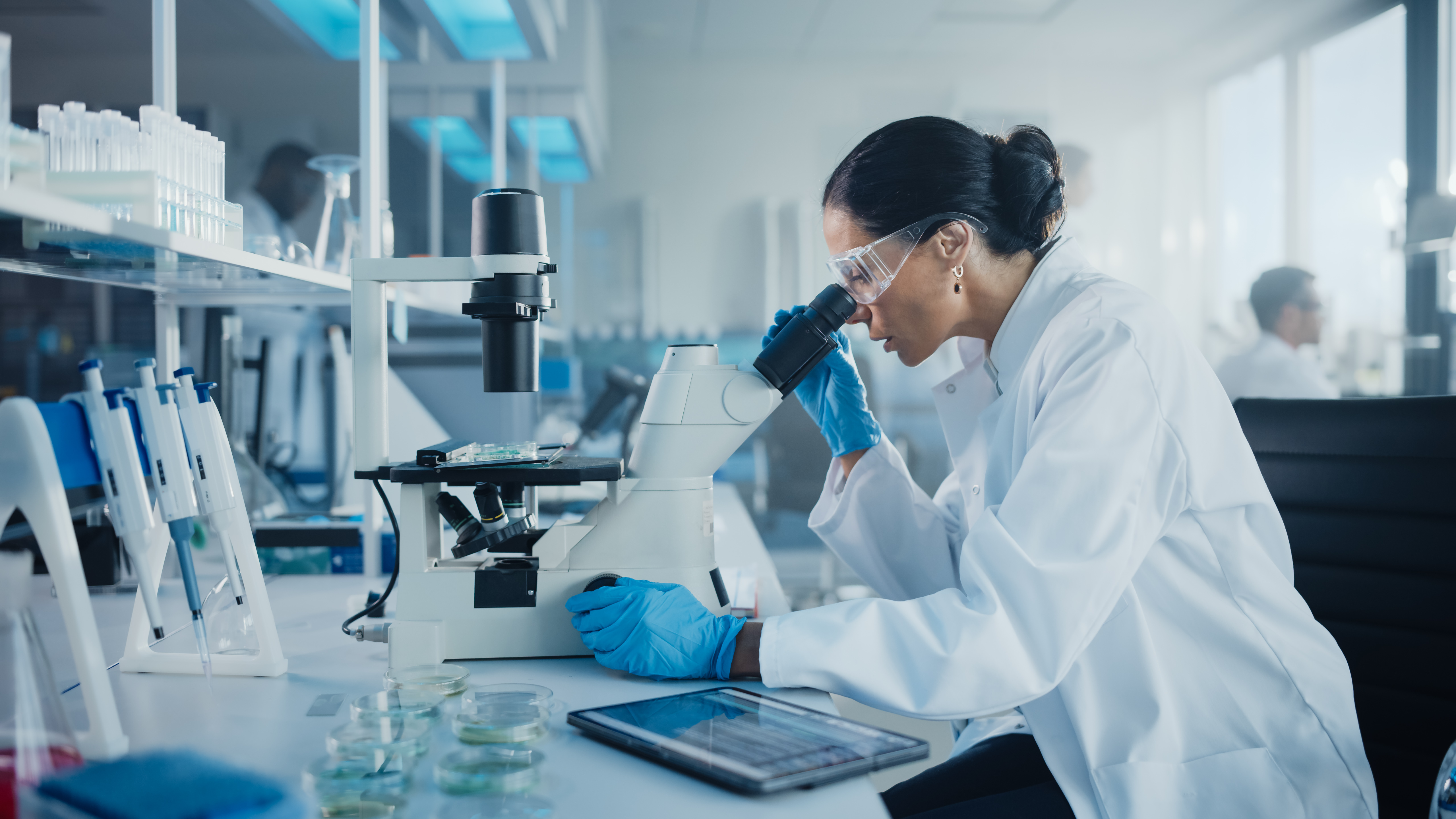 Image of a woman scientist wearing a lab coat and protective gear while looking into a microscope 