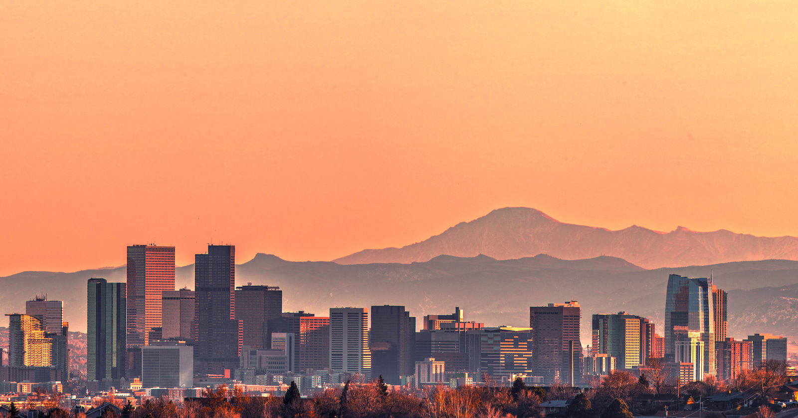 A photograph of the Denver skyline with mountains in the background, against an orange sky.