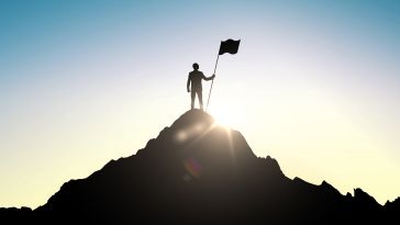 A person stands victoriously on top of a mountain holding a flag in the sunlight..