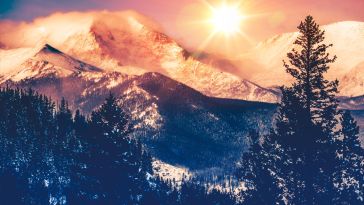 A photograph of the sun shining in the sky above a vista of the Colorado Rocky Mountains.
