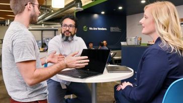 Two men and one woman sitting around a table with a laptop on it, having a discussion. In the background, a blue wall that reads "We are Vendavo" can be seen.