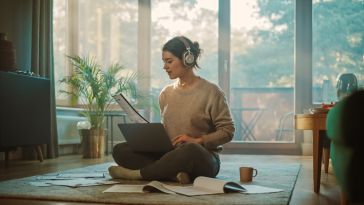 photo of person with headphones on working on laptop while sitting on the floor, with papers strewn about