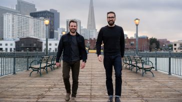 Two Boxes was co-founded by CEO Kyle Bertin (left) and CPO Evan Stalter (right). The two are pictured walking down a wooden pier with buildings in the background.