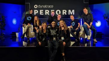 Nine Dynatrace team members sit on a stage with blue backlighting and a sign reading “Dynatrace Perform.”