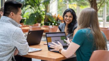 Three Workiva employees sit at outdoor table with laptops, meeting together and smiling