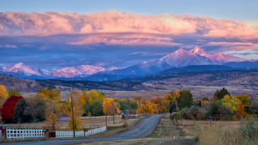 Long's Peak lights up at sunrise as a rural country road leads into the fall trees