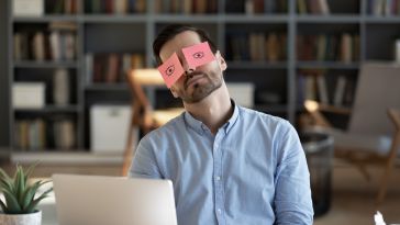 A man sits behind a laptop with sticky notes with eyes drawn on them over his own eyes.